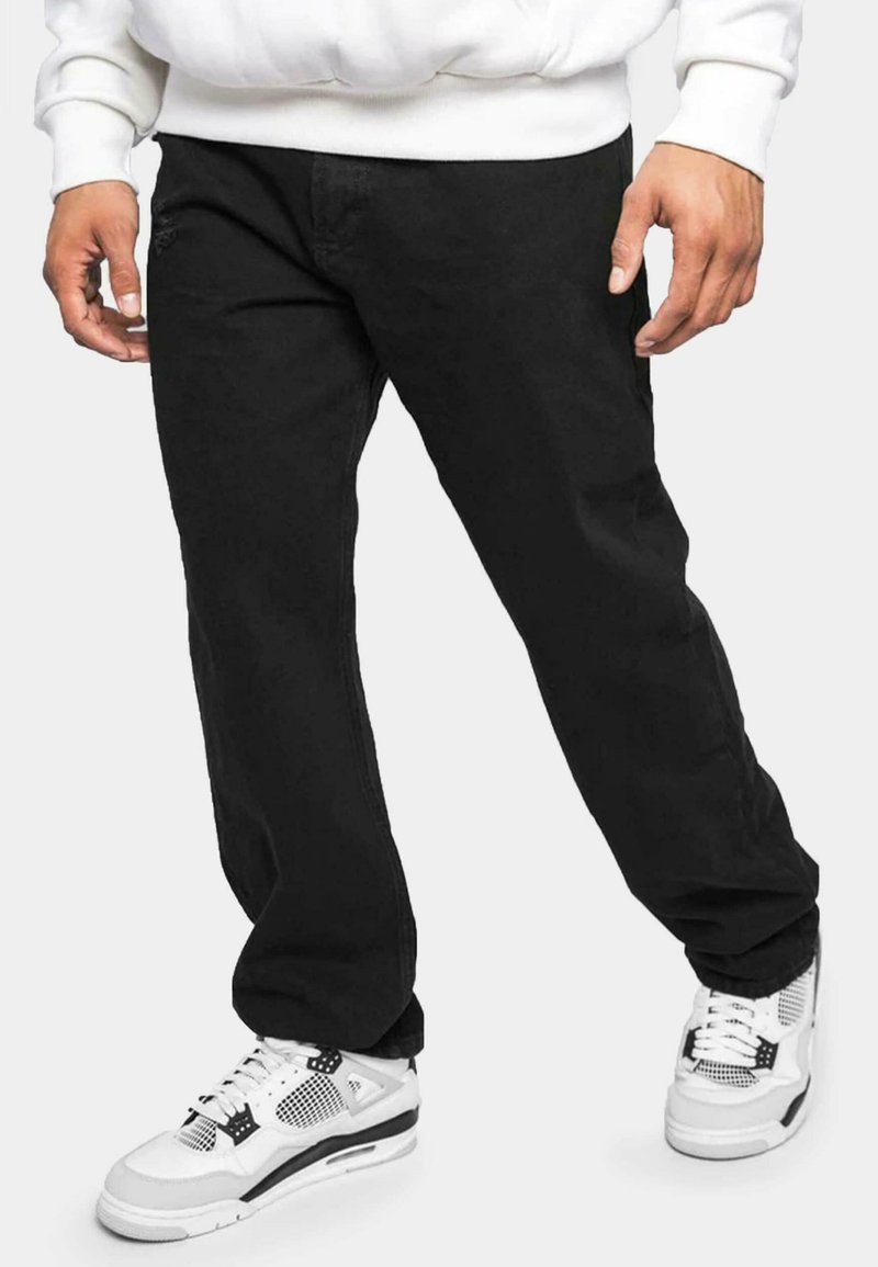 Jeans in denim nero con taglio dritto, dotati di tasche frontali e dettagli di cucitura sottile, abbinati a scarpe sportive bianche e nere.