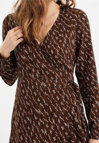 Brun mönstrad omslagsklänning med långa ärmar, med en texturerad tyg och vit sicksackdesign över hela. Knuten midjedetalj synlig.