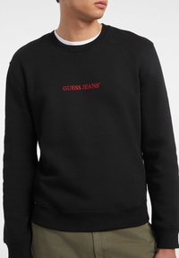 Black sweatshirt made of cotton blend, featuring crew neck and ribbed cuffs. The front displays red embroidered "GUESS JEANS" logo.