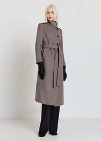 Long manteau marron en mélange de laine avec un col montant, des boutons croisés, une ceinture à la taille et des poches latérales ; accompagné de gants en cuir noir et d'un pantalon noir.