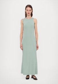 Robe maxi sans manches de couleur vert menthe en tissu lisse, avec un décolleté arrondi et un détail découpé sur une épaule, accompagnée de ballerines noires.