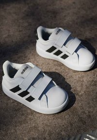 Bijele atletske cipele s tri čičak trake, crnim prugama sa strana, gumenim potplatima i zaobljenim vrhovima prstiju. Detalji s teksturom na trakama.