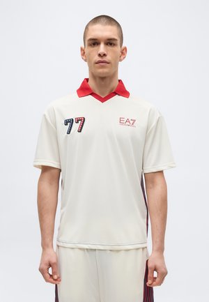 Fiatal férfi áll előre nézve, fehér sportöltözéket viselve piros gallérral, 77-es számmal és EA7 Emporio Armani logóval az ingén.