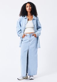 Chaqueta de mezclilla azul clara sobre una camiseta sin mangas blanca y una falda larga azul clara con una abertura frontal, combinada con zapatillas blancas y grises.