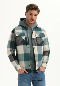 Geruite flanellen overhemd in teal, wit en grijs met twee borstzakken, knoopsluiting en een gestructureerde afwerking, gedragen over een grijze hoodie.
