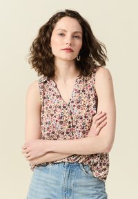 Blusa sin mangas floral con escote en V y detalle de botones, que presenta un fondo claro con patrones de flores multicolores. Combinada con jeans de color azul claro.