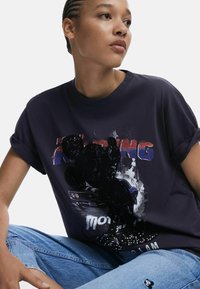 Navyblaue Baumwoll-T-Shirt mit einem grafischen Druck eines Autos, Text und Paillettenakzenten. Aufgerollte Ärmel und lässige Passform.