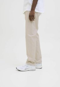 Persoană purtând pantaloni bej și adidași albi, stând cu fața spre lateral, pe un fundal alb simplu.