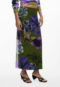 Maxi rok van groene stof met grote paarse en witte bloemenpatronen. Gecombineerd met zwarte sandalen metbandjes, wat het elegante ontwerp benadrukt.