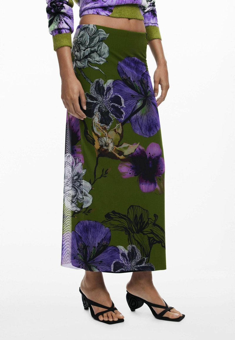 Maxi rok van groene stof met grote paarse en witte bloemenpatronen. Gecombineerd met zwarte sandalen metbandjes, wat het elegante ontwerp benadrukt.