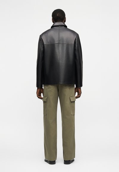 Άνδρας με μαύρο δερμάτινο μπουφάν και χακί παντελόνι cargo στέκεται γυρισμένος με την πλάτη σε απλό λευκό φόντο.