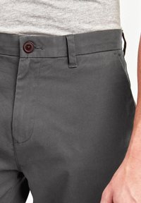 Chinos gris en tissu lisse, avec une taille standard, un bouton marron et une poche latérale, mettant en valeur une coupe ajustée.