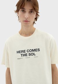 Camiseta de manga corta color crema con el texto en negrita "AQUÍ VIENE EL SOL" en negro, y detalles impresos adicionales en la parte inferior. Material de algodón suave.