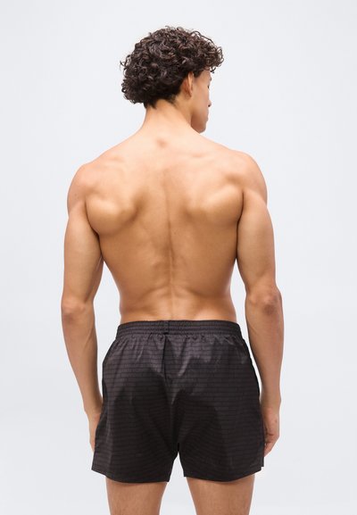 Μυώδης άνδρας με σγουρά μαλλιά, εμφανιζόμενος από πίσω, φοράει μαύρο αθλητικό σορτς σε απλό φωτεινό φόντο.