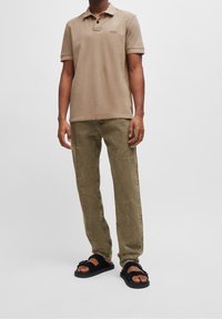 Beige polo shirt met korte mouwen en knoopsluiting, gecombineerd met olijfgroene textuurbroek en zwarte slipper sandalen.