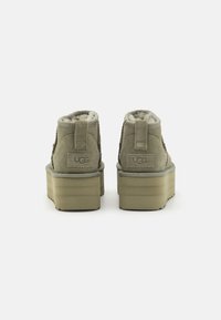 Szare zamszowe botki za kostkę z puszystą wyściółką, podwyższoną platformą i logo UGG z tyłu. Teksturowane boki i wzmocniony cokół.