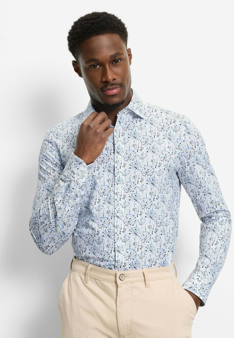 Mann, der den Kragen seiner hellblauen Bluse mit floralem Muster und beiger Hose richtet, steht vor einem schlichten weißen Hintergrund.