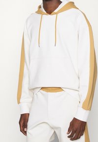 Vit huvtröja med gula accenter, med en framficka, snoddar och ribbade ärmslut. Mjukt bomullsmaterial. Matcher med shorts i samma stil.