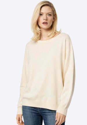 Femme blonde portant un pull crème ample et un jean bleu, debout devant un fond clair uni, face à la caméra.