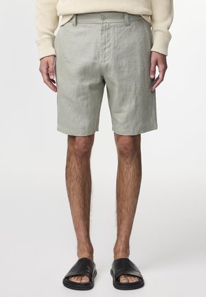 Hellgrüne Leinen-Shorts mit Knopfverschluss, zwei seitlichen Taschen und lockerer Passform, kombiniert mit schwarzen Riemensandalen.