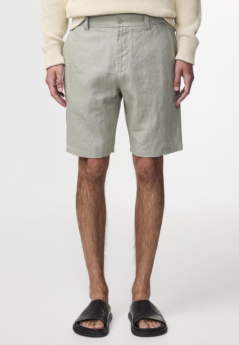 Hellgrüne Leinen-Shorts mit Knopfverschluss, zwei seitlichen Taschen und lockerer Passform, kombiniert mit schwarzen Riemensandalen.
