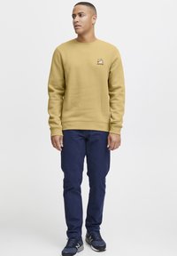 Yellow sweatshirt with round neckline and ribbed cuffs, featuring a small logo on the chest. Paired with dark blue pants and athletic shoes.