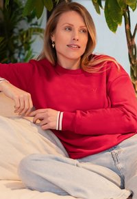 Rode sweatshirt met witte gestreepte manchetten, gemaakt van zachte stof. Ggedragen met lichte blauwe relaxed-fit jeans. Achtergrond met groene planten.