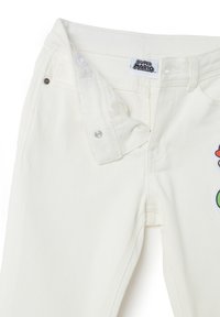 Pantalones de mezclilla blancos con cierre de cremallera y broche, que cuentan con una etiqueta etiquetada como "Super Mario" y un diseño bordado colorido.