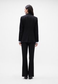 Zwarte op maat gemaakte blazer met een gladde textuur, gecombineerd met bijpassende broek. Opvallende details zijn de knopen op de mouwen van de blazer.
