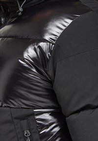 Svart uppblåsbar jacka med glansiga och matta tygtexturer, en ficka med dragkedja och en justerbar huva med snörningar.