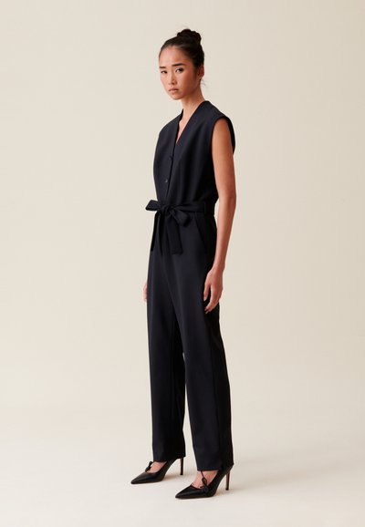 Tutina nera con vita cinchata e maniche corte, caratterizzata da una chiusura con bottoni, vestibilità sartoriale e pantaloni a gamba larga. Indossata con tacchi neri.
