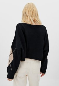 Suéter negro corto con un corte holgado y mangas grandes, con un patrón de rombos beige en la manga derecha. Tela de punto texturizada.