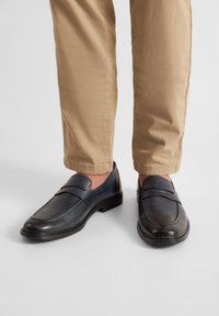 Tmavomodré kožené mokasíny s hladkou textúrou, ozdobené dvoma pásikmi cez prednú časť, skombinované s béžovými nohavicami.