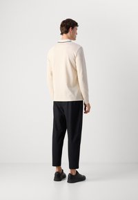 Mann iført en kremfarget polo-skjorte med lange ermer, svarte cropped bukser og svarte sneakers, som står med ryggen til mot en hvit bakgrunn.
