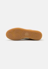 Semelle de sneaker présentant une surface en caoutchouc texturé de couleur brun clair, avec un logo en relief et un motif conçu pour une meilleure adhérence.