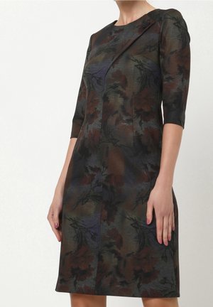 Mujer que lleva un vestido ajustado hasta la rodilla con mangas hasta el codo y un patrón floral oscuro en tonos apagados de verde, marrón y azul marino.