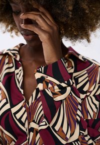 Person with curly hair touching their face, wearing a multicolored shirt with bold abstract patterns in black, cream, maroon, brown, and purple.