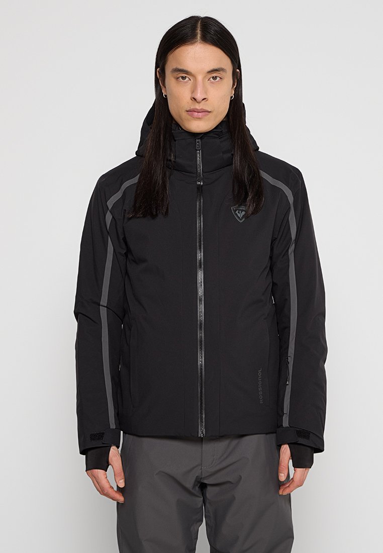 Person mit langen dunklen Haaren, die eine schwarze Zip-Jacke mit grauen reflektierenden Streifen und graue Hose trägt, steht vor einem schlichten Hintergrund.