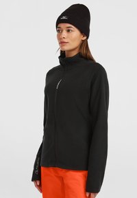 Svart fleecejacka med hög krage, dragkedja och Polartec-logotyp på ärmarna. Modell bär orange byxor och en svart stickad basker.