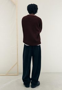 Vinröd stickad tröja med ribbad textur, tillsammans med lösa svarta kordflorsebyxor. Figuren står med armarna i kors, med ryggen vänd mot oss.