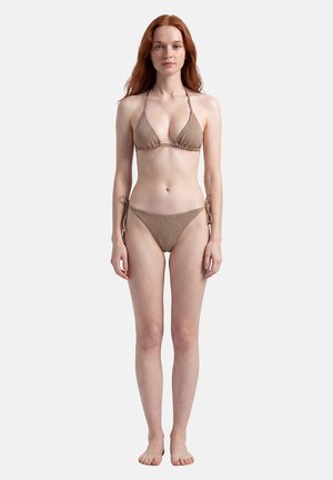 Donna con lunghi capelli rossi, in piedi a piedi nudi, indossa un bikini marrone con fiocchi sui lati su sfondo chiaro e uniforme.