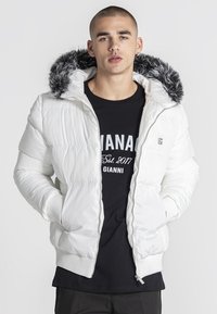 Bijela podstavljena jakna s kapuljačom obloženom umjetnim krznom, koja sadrži prednji zip, rebraste manžete i glatku teksturu. Nosena preko crne majice.