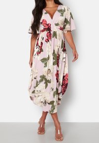 Femme portant une robe cache-coeur rose avec un imprimé floral rouge et crème, des manches flottantes et des sandales à talons avec des brides transparentes, se tenant devant un fond blanc.