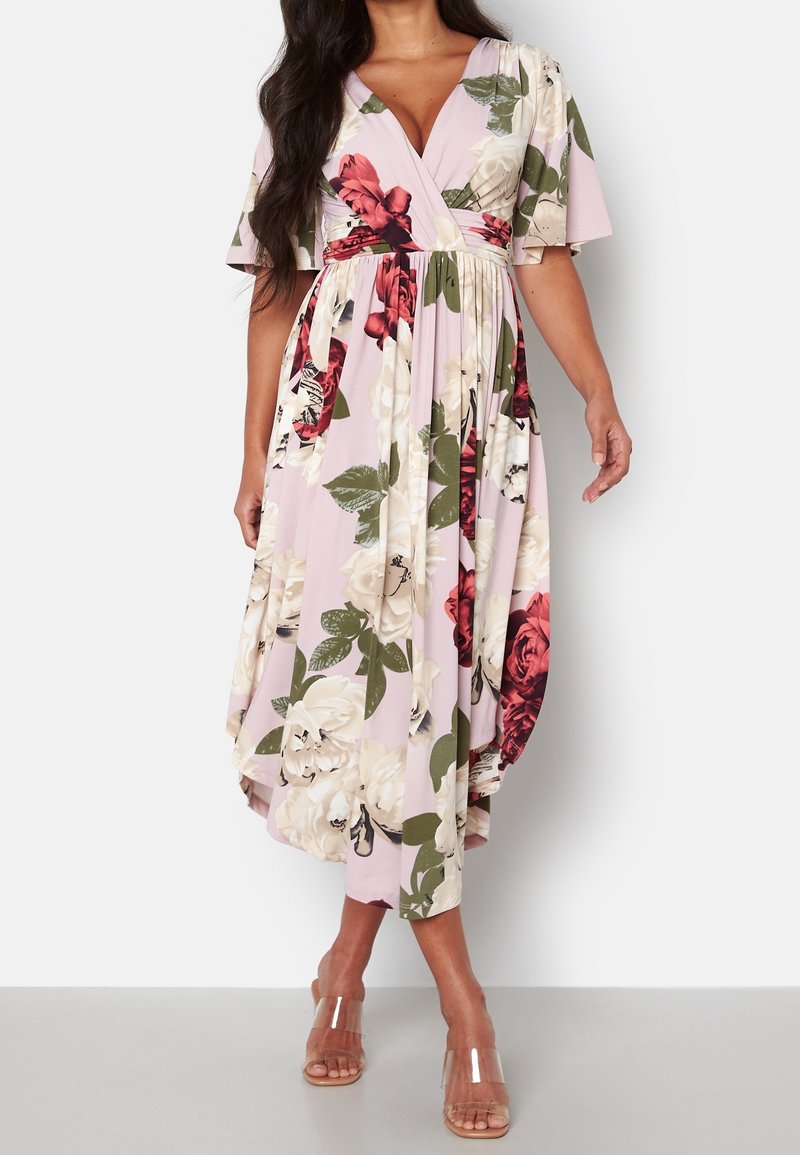 Femme portant une robe cache-coeur rose avec un imprimé floral rouge et crème, des manches flottantes et des sandales à talons avec des brides transparentes, se tenant devant un fond blanc.