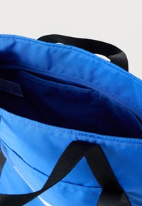 Τσάντα ώμου από μπλε ύφασμα με φαρδιά άνοιγμα, διαθέτει μαύρες χειρολαβές και εσωτερική τσέπη. Λείος υφασμάτινος σχεδιασμός και στιβαρή κατασκευή.