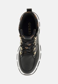 Zapatilla de cuero negra con herrajes dorados, patrón de logo marrón en el cuello interior, suela media blanca y detalles texturizados en la parte superior.