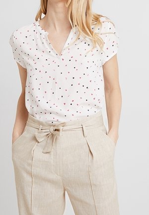 Kvinde iført en hvid kortærmet bluse med små sorte og lyserøde blomsterprint og beige bukser med høj talje og bindebælte.