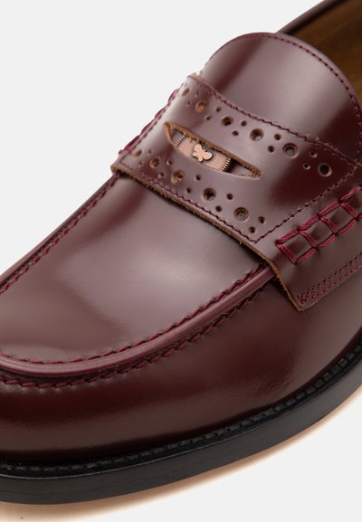 Μπορντό δερμάτινο loafer με διακοσμητικές τρύπες, ραμμένες λεπτομέρειες και μικρό μεταλλικό έμβλημα πεταλούδας στο λουράκι.