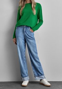 Pull en maille verte à col rond, associé à un jean large bleu clair. Baskets blanches avec semelles beiges. Tissu texturé visible.