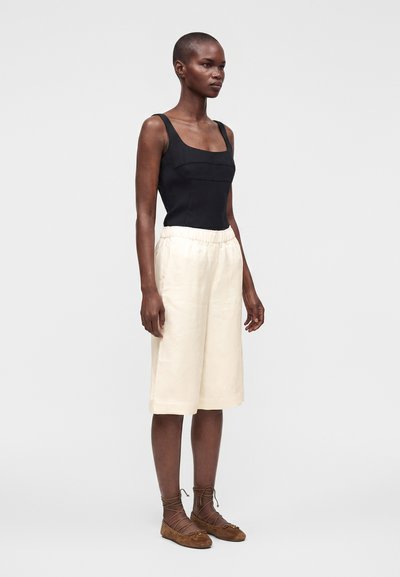 Femme portant un haut noir sans manches, un short ample crème longueur genou et des chaussures plates marron à lacets, debout devant un fond clair.